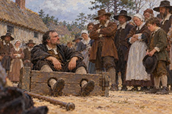 A bareheaded man sits on sandy ground with his ankles locked in wooden stocks, glaring up at a half circle of townspeople in plain wool clothes before a small thatched town house.