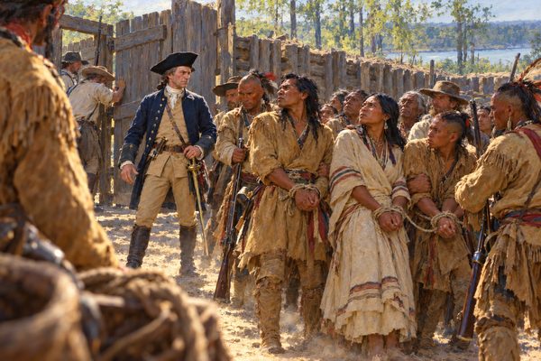 Inside a log fort's gate, armed men bind the wrists of a group of Indian men and women while a coated officer with a sword stands watching; the captives look back at him, steady and angry, in soft spring light.