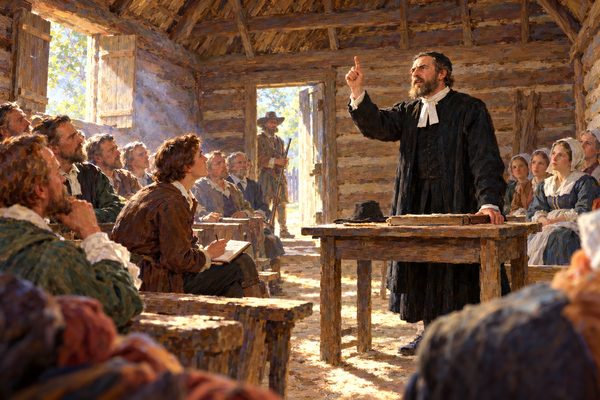 A man in black stands at a plain wooden table in a small log meeting room, one hand raised, while townspeople sit on rough benches and a young man writes in a notebook, in shafts of daylight.