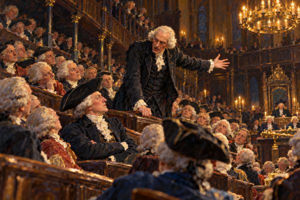 In a packed, candle-lit chamber of tiered benches a gaunt man in a white wig stands gripping the bench before him, one arm flung out, looking down at a seated man with folded arms, as rows of wigged members turn to watch.