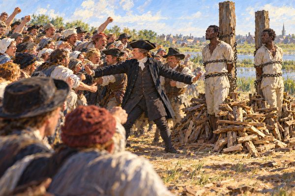 A sheriff with arms spread holds back a packed, shouting crowd in front of two tall stakes where two chained men stand upright, wood piled at their knees, on open ground under a spring sky.