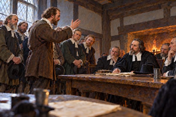 In a plain timber room, a farmer in a brown coat raises one hand in argument at the end of a table, facing a grave man in black seated behind it with other magistrates, while his neighbors stand behind him.